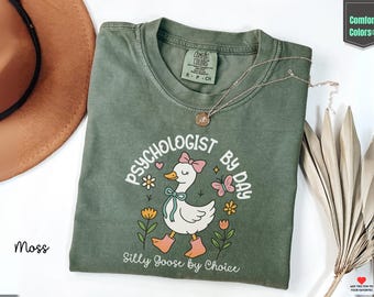 May include: A moss green Comfort Colors t-shirt with the text "PSYCHOLOGIST BY DAY" and "Silly Goose by Choice". The shirt features a cartoon goose wearing a bow, boots, and a bow tie. A gold necklace is draped across the shirt.