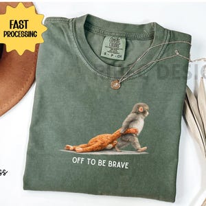 May include: Moss green t-shirt with a graphic of a monkey pulling a stuffed animal, with the text "OFF TO BE BRAVE". The shirt has a crew neck and short sleeves. A gold necklace is draped on the shirt.