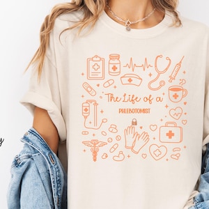 May include: Ivory t-shirt featuring an orange medical-themed design. The design includes medical symbols, a heartbeat line, and the text "The Life of a Phlebotomist".