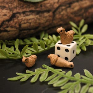 Puede incluir: Miniaturas de ardillas marrones, una encima de un dado blanco con puntos negros y otras dos cerca. Las ardillas están rodeadas de follaje verde, creando una escena caprichosa inspirada en la naturaleza. Las figuras son detalladas y parecen hechas a mano.