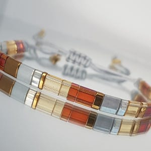 May include: A colorful bracelet with a sliding knot closure. The bracelet features rectangular beads in shades of gold, red, brown, and silver. The bracelet is displayed on a reflective surface.