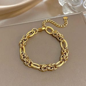 May include: A gold bracelet featuring a mix of chain link styles. The bracelet has a combination of oval and woven links, with an adjustable chain for sizing. The jewellery is displayed on a neutral surface.