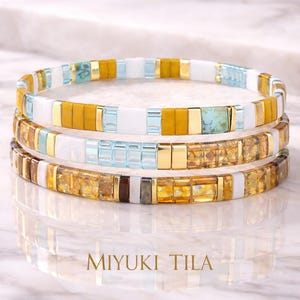 May include: Three beaded bracelets with a mosaic tile design. The bracelets feature a mix of colors, including gold, white, yellow, and blue. The text "MIYUKI TILA" is displayed at the bottom of the image.