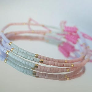 May include: Stack of beaded bracelets in pastel colors. The bracelets are made of small, rectangular beads in shades of pink, blue, and green, with gold-colored accents. The bracelets are delicate and feminine.
