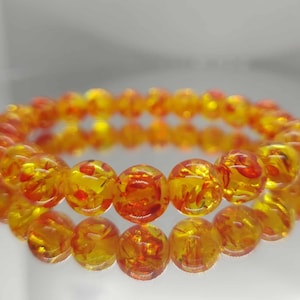 May include: A bracelet made of translucent, round beads in yellow and orange hues. The beads have a speckled, marbled effect. The bracelet is displayed on a reflective surface. The beads are approximately 1 cm in diameter.