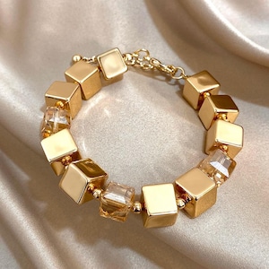 May include: Gold-coloured bracelet with alternating square and faceted crystal beads. The bracelet has a chain clasp and small gold beads between the larger beads. The cubes have a polished finish, reflecting light. The jewellery is displayed on a satin surface.