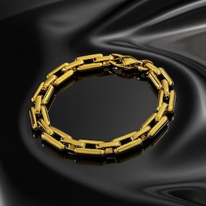 Op de afbeelding: Een goudkleurige armband met een rechthoekig schakelontwerp. De glanzende armband wordt gepresenteerd op een zwart oppervlak. De sluiting is zichtbaar en de algehele esthetiek is er een van luxe en elegantie.