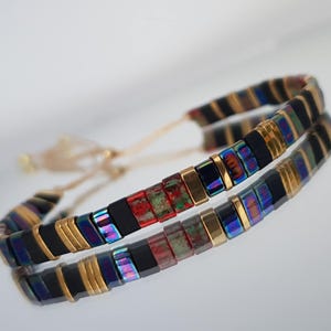 May include: A beaded bracelet featuring a mix of black, gold, and iridescent rectangular beads. The bracelet is strung on a beige cord and is reflected in a shiny surface. The beads have a colorful, oil-slick appearance.