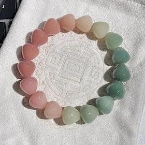 May include: A bracelet made of heart-shaped beads in a gradient of colours, from pink to green. The beads are arranged in a circular pattern, resting on a white, textured surface.