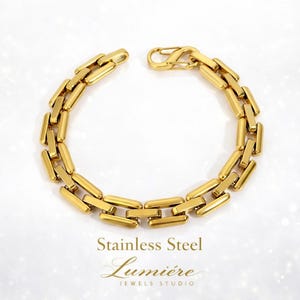 Pode incluir: Uma pulseira dourada de aço inoxidável com um design de elos retangulares. A pulseira está aberta, mostrando o mecanismo de fecho. O texto "Stainless Steel Lumière Jewels Studio" é visível abaixo.