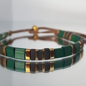 Puede incluir: Pulsera hecha a mano con un patrón repetido de cuentas rectangulares verde oscuro, doradas y marrón oscuro. La pulsera está ensartada en un cordón marrón con nudos corredizos ajustables. Diseño sencillo y elegante.