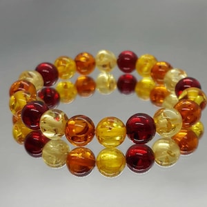 May include: A bracelet made of amber beads in various colours, including yellow, orange, and red. The beads are spherical and arranged in a circular pattern. The bracelet is shown on a reflective surface.