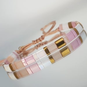 Könnte beinhalten: Zwei passende Armbänder mit einem Mosaikfliesen-Design. Die Fliesen sind weiß, rosa und goldfarben. Die Armbänder sind durch eine pfirsichfarbene Kordel mit verstellbarem Knotenverschluss verbunden. Die Armbänder werden auf einer reflektierenden Oberfläche präsentiert.