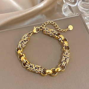 Op de afbeelding: Goudkleurige armband met een Byzantijns kettingontwerp. De armband heeft een karabijnsluiting en een verstelbare ketting. De schakels zijn ingewikkeld geweven, waardoor een accessoire met textuur ontstaat. De armband wordt gepresenteerd op een neutrale ondergrond.