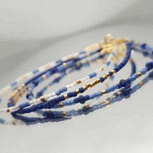以下が含まれることがあります： 青、白、金色のビーズブレスレットの束。 ブレスレットは、小さな丸いビーズで作られています。 金色のビーズがデザインにエレガントさを加えています。 ブレスレットは反射する表面に表示されています。