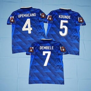 Può includere: Tre maglie da calcio blu con numeri e nomi dei giocatori bianchi. Le maglie presentano i nomi "UPAMECANO", "KOUNDE" e "DEMBELE" con i numeri 4, 5 e 7. Ogni maglia ha un logo sulle maniche.