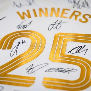 May include: White jersey with gold lettering and the number 25. The word "WINNERS" is arched above the number. Numerous signatures in black ink are scattered across the jersey. A logo with a lion and soccer ball is in the corner.