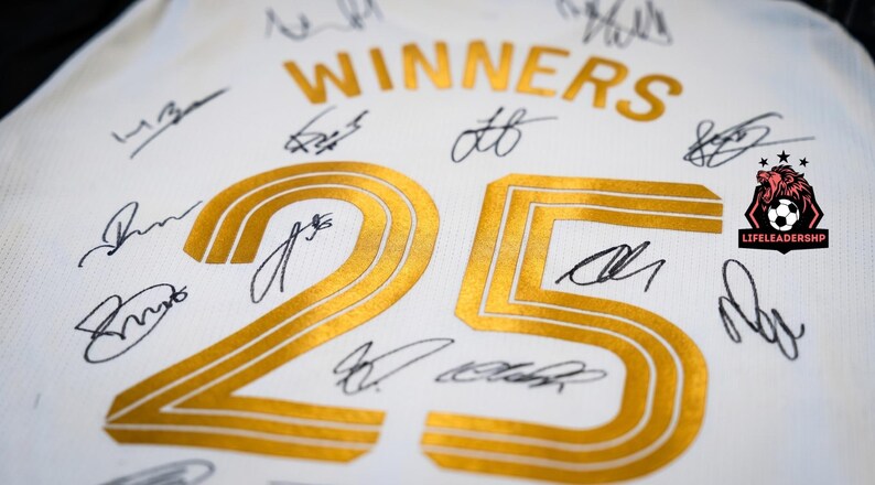 May include: White jersey with gold lettering and the number 25. The word "WINNERS" is arched above the number. Numerous signatures in black ink are scattered across the jersey. A logo with a lion and soccer ball is in the corner.