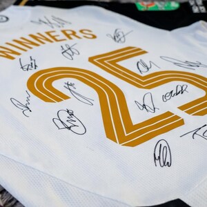 May include: White and black soccer jersey with gold accents and player signatures. The jersey features the word "WINNERS" above a large gold number "25". The jersey is likely a team souvenir or collectible.