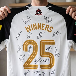May include: White soccer jersey with black sleeves, featuring the word "WINNERS" and the number "25" in gold. The jersey is covered in signatures and includes logos on the sleeves. The jersey is held up by a person.
