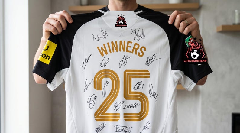 May include: White soccer jersey with black sleeves, featuring the word "WINNERS" and the number "25" in gold. The jersey is covered in signatures and includes logos on the sleeves. The jersey is held up by a person.