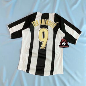May include: A black and white striped football shirt with the name "IBRAHIMOVIC" and the number "9" in gold. The shirt has a white collar and sleeves. A logo is visible on the lower right corner.