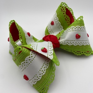 May include: A large, decorative hair bow featuring a vibrant green and white design with red accents. The bow is adorned with small, red strawberry embellishments and a delicate lace trim. The center of the bow is a bright red color.