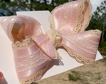 Puede incluir: Un lazo decorativo grande de satén rosa pálido, adornado con un ribete de encaje color crema. El lazo presenta un diseño en capas con un nudo central y se presenta sobre una superficie blanca, lo que sugiere un accesorio para el cabello o una decoración de regalo.