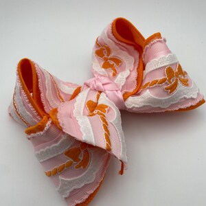 Peut inclure: Un nœud décoratif pour cheveux composé de rubans roses superposés et de dentelle blanche. Le motif présente des nœuds orange brodés et des détails en forme de corde. Les bords sont finis par un liseré orange vif et le centre est noué en rose.