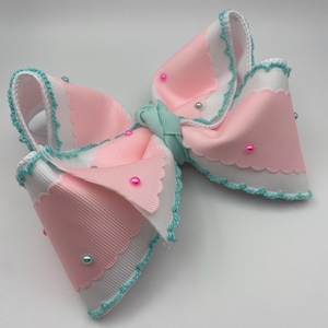 Op de afbeelding: Een haarstrik gemaakt van gelaagd wit en lichtroze grosgrain lint met geschulpte randen. De randen zijn afgewerkt met een dunne, groenblauwe gehaakte boord. De strik is versierd met kleine, verspreide roze en lichtblauwe ronde kraaltjes.