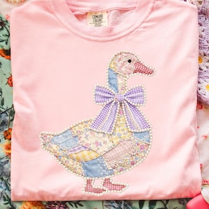 Op de afbeelding: Een lichtroze Comfort Colors t-shirt met een patchwork ganzenontwerp en een paarse ruitjesstrik. De gans is gemaakt van diverse bloemen- en patroonstoffen, met parelrand. Het shirt wordt gepresenteerd op een stof met bloemenpatroon.