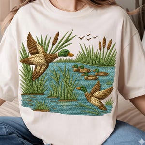 May include: Cream-colored t-shirt featuring a detailed illustration of ducks in a pond. The design includes ducks in flight and swimming, surrounded by reeds and cattails. The artwork uses shades of brown, green, and blue.