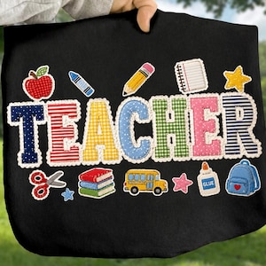 May include: A black t-shirt featuring the word TEACHER in large, multi-colored patterned letters. Surrounding the text are graphic icons including an apple, pencil, notebook, star, scissors, stack of books, school bus, glue, and a backpack.