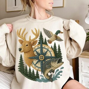 May include: Beige sweatshirt featuring a nature-themed graphic. The design includes a deer, duck, fish, compass, and pine trees. The artwork uses shades of brown, green, and blue. The sweatshirt is a casual garment.