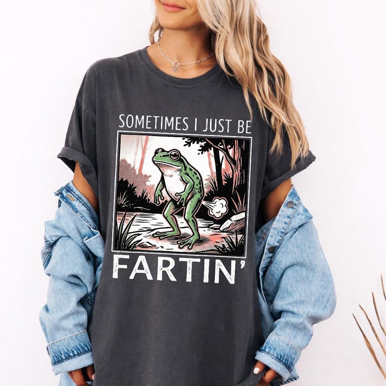 May include: Dark gray t-shirt with the text "SOMETIMES I JUST BE FARTIN'" and a graphic of a green frog in a forest scene. The frog is depicted mid-fart, with a cloud of gas emanating from its rear.