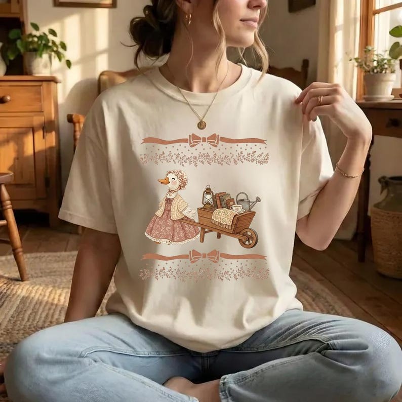 Puede incluir: Camiseta color crema con una ilustraci&oacute;n caprichosa de un pato con gorro y vestido empujando una carretilla llena de libros, una linterna y una regadera. El dise&ntilde;o incluye lazos decorativos y elementos florales en tonos marrones.