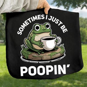 May include: Black tote bag featuring a cartoon frog holding a coffee cup. The frog is green with white accents and sits on a mossy rock. The text "SOMETIMES I JUST BE POOPIN'" is printed in white around the frog.