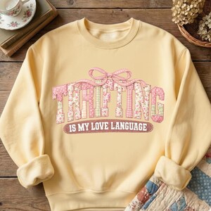Puede incluir: Una sudadera amarilla clara con la palabra "THRIFTING" en letras de patchwork rosa. El texto "IS MY LOVE LANGUAGE" est&aacute; debajo. La sudadera tiene mangas largas y cuello redondo.