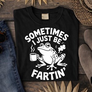 May include: Black t-shirt with a white graphic of a frog holding a coffee cup, with the text "SOMETIMES I JUST BE FARTIN'". The shirt is folded on a wooden surface, with a pair of blue jeans and sunglasses visible.