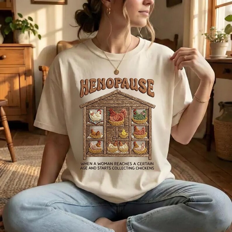 Puede incluir: Camiseta color crema con la palabra "HENOPAUSE" en letras marrones sobre un gr&aacute;fico de un gallinero. El gallinero contiene varias gallinas en cestas. El texto debajo del gallinero dice: "CUANDO UNA MUJER ALCANZA CIERTA EDAD Y COMIENZA A COLECCIONAR POLLOS."