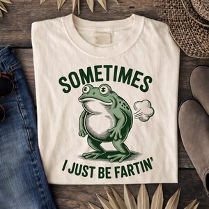 May include: Cream-colored t-shirt with a green frog illustration and the text "SOMETIMES I JUST BE FARTIN'". The frog is depicted with a cloud of gas. The shirt is folded on a wooden surface.