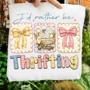 Puede incluir: Bolso tote blanco con el texto "I'd rather be Thrifting". El dise&ntilde;o incluye ilustraciones de un lazo amarillo, un carrito de compras lleno de art&iacute;culos y un lazo rosa. La palabra "Thrifting" est&aacute; en letras coloridas y estampadas.
