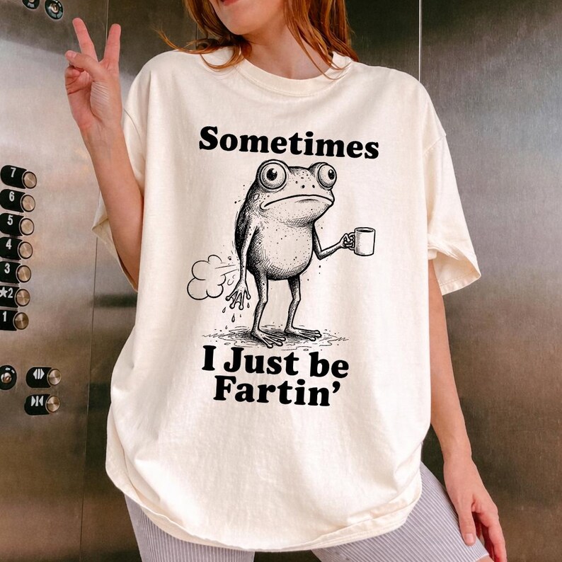 May include: Cream-colored t-shirt with the text "Sometimes I Just be Fartin'" above a cartoon frog holding a mug. The frog is drawn in black and white, with a cloud of gas emanating from behind it.