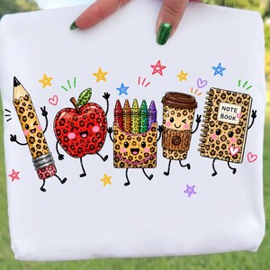 May include: A white t-shirt featuring a colorful graphic of five anthropomorphic school supplies with leopard print patterns: a pencil, an apple, a box of crayons, a coffee cup, and a notebook labeled NOTE BOOK, surrounded by small stars.