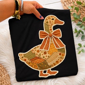 May include: A black t-shirt featuring a large appliqué of a goose. The goose is constructed from various fabric patches in shades of orange, brown, and green with leaf patterns and gingham checks. A large bow is at the neck, and pearl trim outlines the goose.