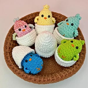 May include: Five crocheted chicks in white eggshells, sitting in a brown crocheted nest. The chicks are yellow, pink, blue, green, and light blue.