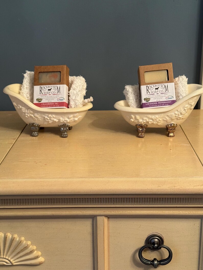 May include: Two bars of handmade soap in small white porcelain tubs with feet. The soap on the left is labeled "Cranberry Fig" and the soap on the right is labeled "Lavender Peppermint". Both soaps are from Rockbottom Soap Co.