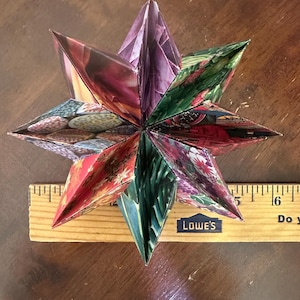 Pode incluir: Uma estrela de origami multicolorida com várias seções de papel padronizado. A estrela, com aproximadamente 15 cm de diâmetro, repousa sobre uma régua de madeira. As cores da estrela incluem tons de roxo, verde, vermelho e azul.