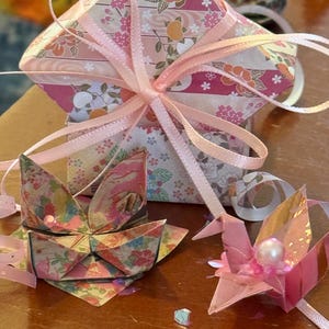 May include: Origami paper crafts in shades of pink, peach, and floral patterns. A square gift box tied with a pink ribbon, a folded boat, and a crane with a pearl accent are visible.