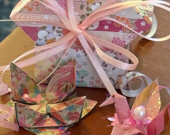 May include: Origami paper crafts in shades of pink, peach, and floral patterns. A square gift box tied with a pink ribbon, a folded boat, and a crane with a pearl accent are visible.